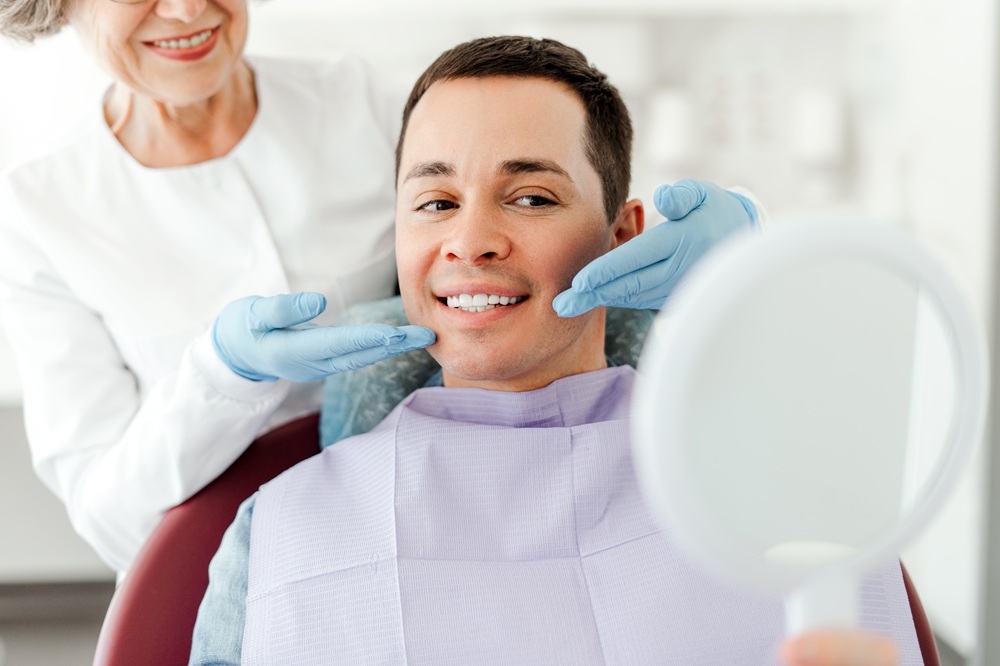 A smiling man wearing a lavender dental bib holds a mirror while a dentist in gloves points to his teeth, showcasing his bright smile. - Gramercy Dental Group