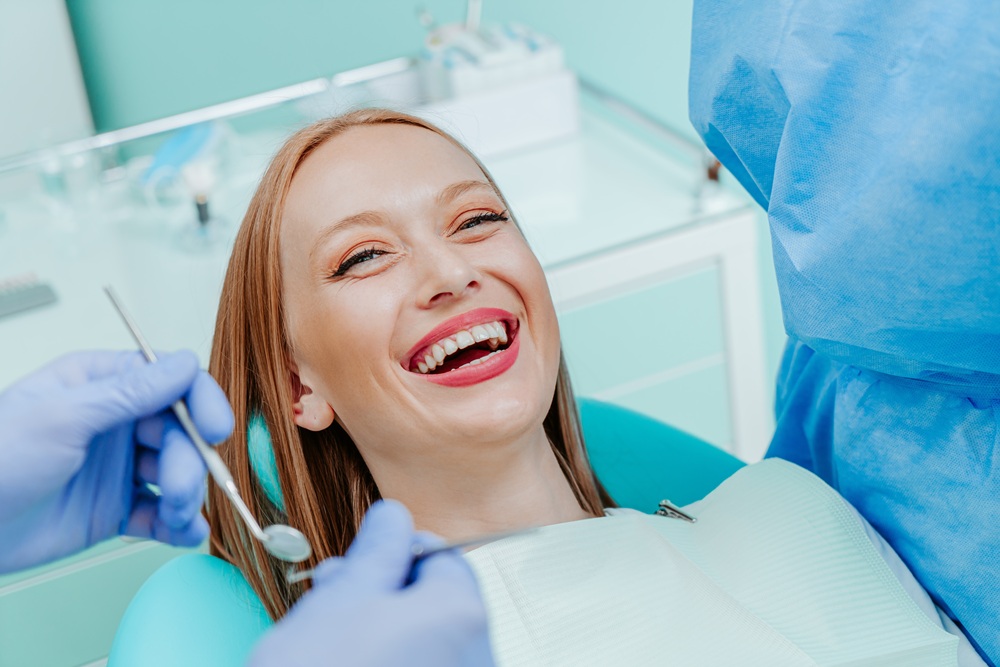 A cheerful woman with long red hair laughs while seated in a dental chair as two gloved hands hold dental instruments – Dentist by Union Square