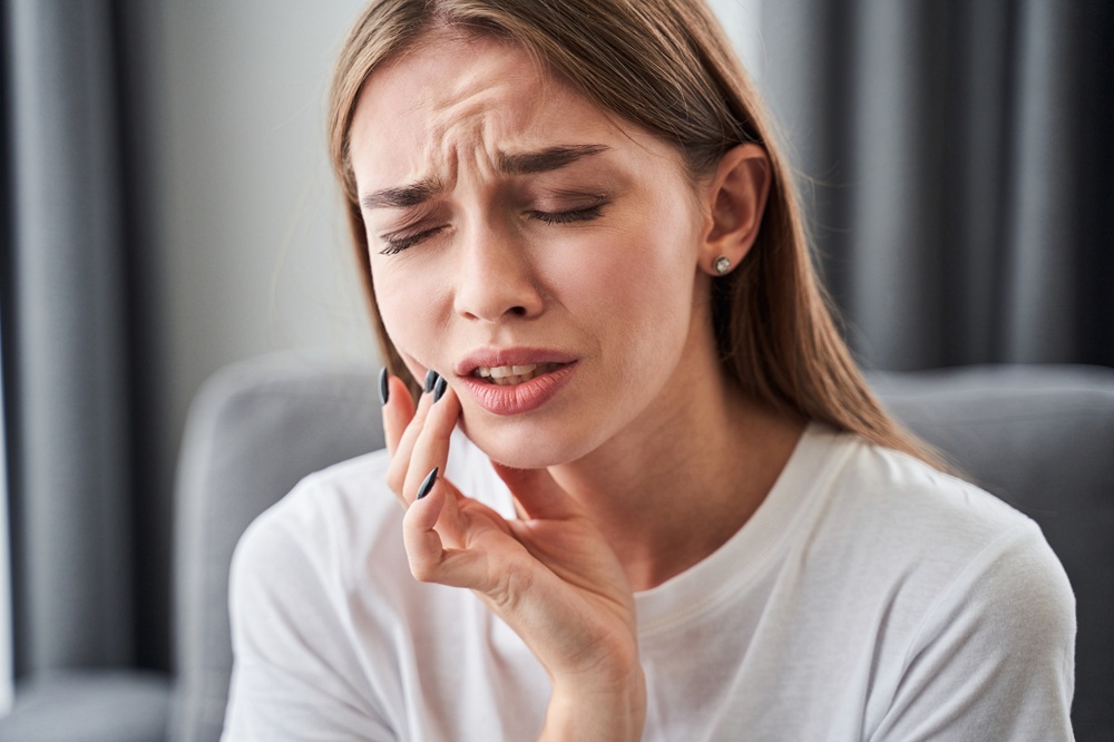 Woman Holding Jaw in Pain – Dental Emergencies A young woman in a white shirt touches her cheek with a pained expression, suggesting tooth or jaw discomfort – Dental Emergencies
