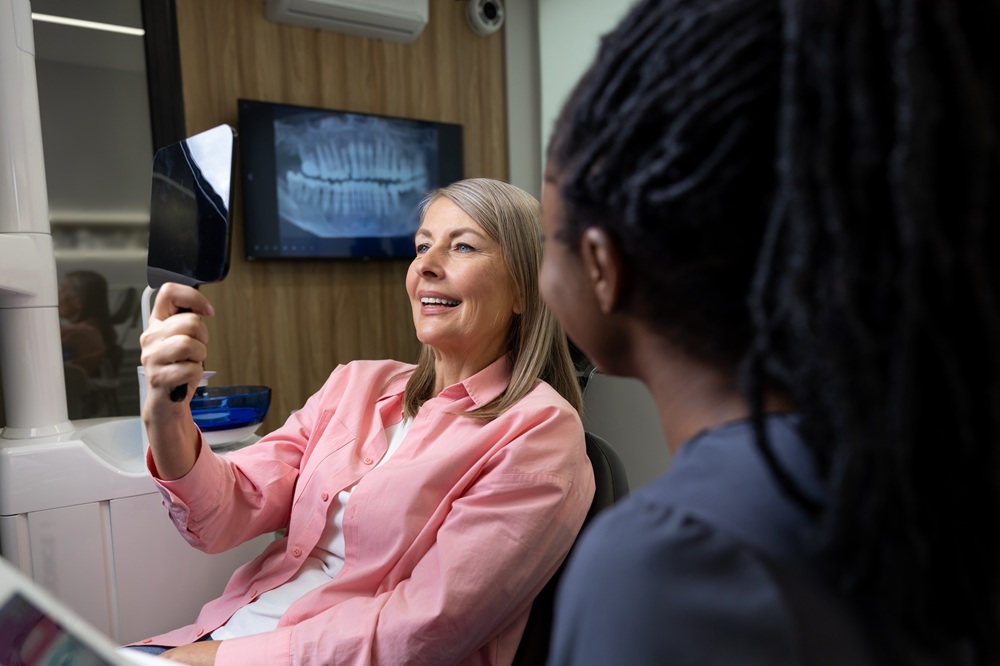 Patient Smiling in Dental Office – Chelsea Dental Implants A woman in a pink shirt smiles while holding a mirror in a dental office, with a panoramic dental X-ray visible on the screen behind her – Chelsea Dental Implants