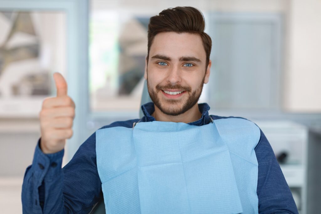 A smiling man with a blue dental bib sits in a modern dental clinic chair, giving a thumbs-up gesture, looking satisfied after his appointment. - Gramercy Dental Group