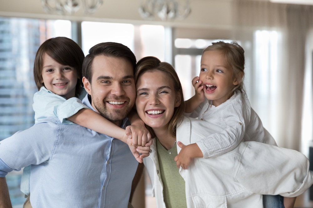 A happy family of four smiles brightly indoors, with both children playfully riding on their parents’ backs – Emergency Dentist East Village