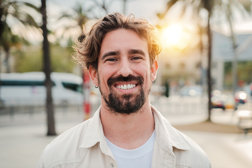 A cheerful man with wavy hair and a beard smiles warmly while standing outside near palm trees and city traffic at sunset – Emergency Dentist East Village