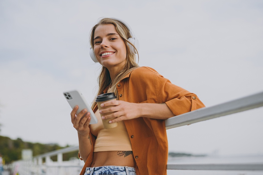 A cheerful young woman wearing headphones and an orange shirt leans on a railing, holding a smartphone and a coffee cup while smiling at the camera. - Gramercy Dental Group