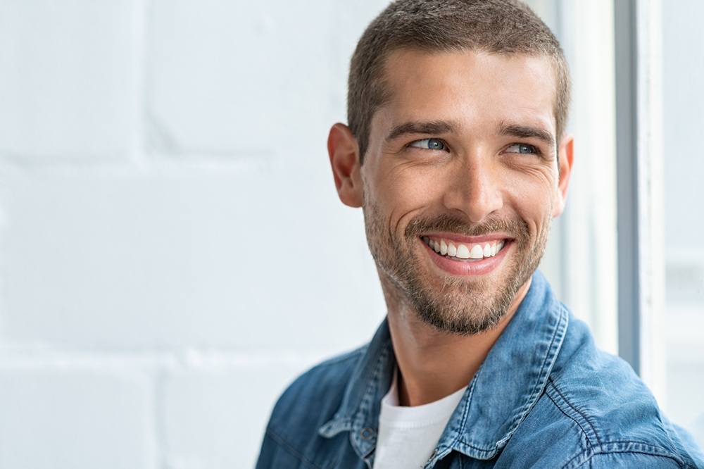 A man with short hair and a trimmed beard smiles brightly while looking to the side, wearing a denim shirt near a sunlit window – Chelsea Dentist