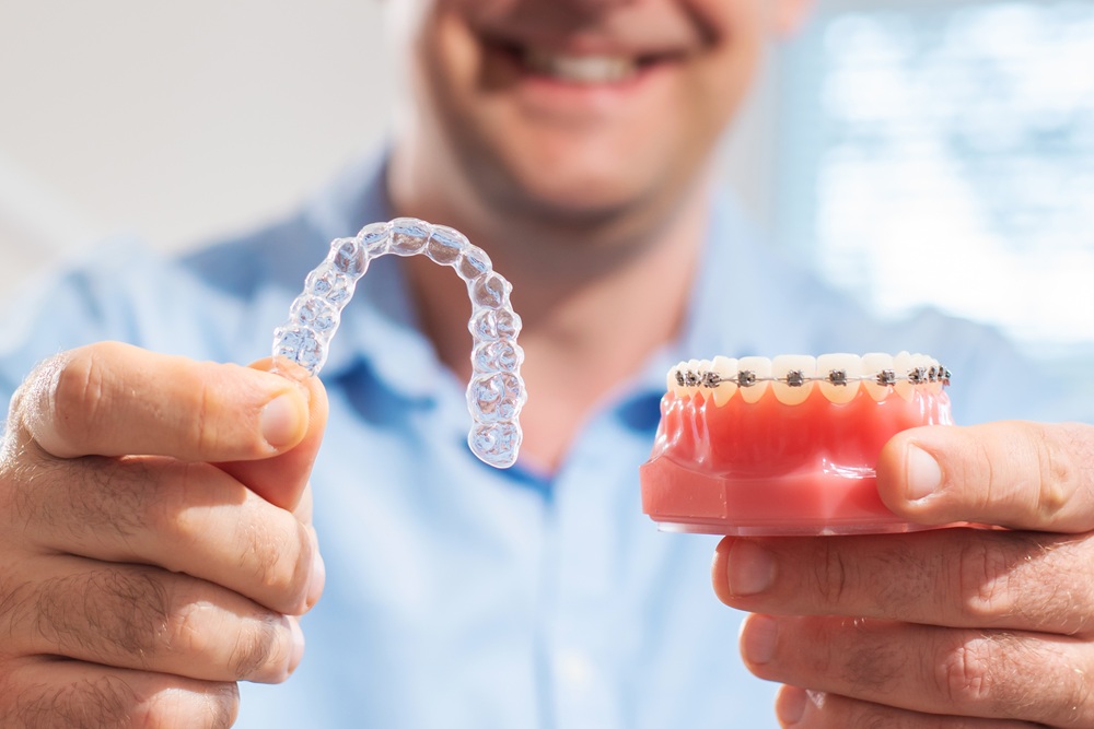 Comparing Invisalign Aligner with Braces Model – Invisalign A person holding a clear Invisalign aligner in one hand and a dental model with traditional metal braces in the other, demonstrating orthodontic options – Invisalign