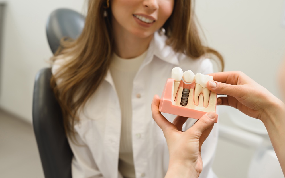 Patient Consultation with Dental Implant Model – Chelsea Dental Implants A dental professional shows a model of a dental implant to a smiling female patient during a consultation – Chelsea Dental Implants