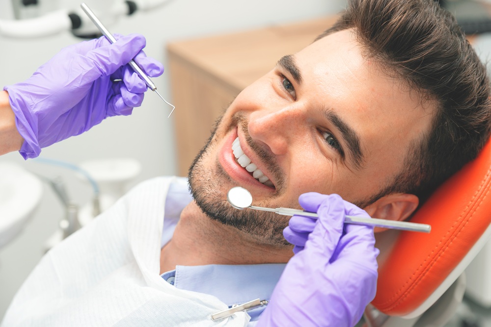 A cheerful man reclines in a dental chair while a dentist wearing purple gloves holds dental tools near his smile – Dentist East Village
