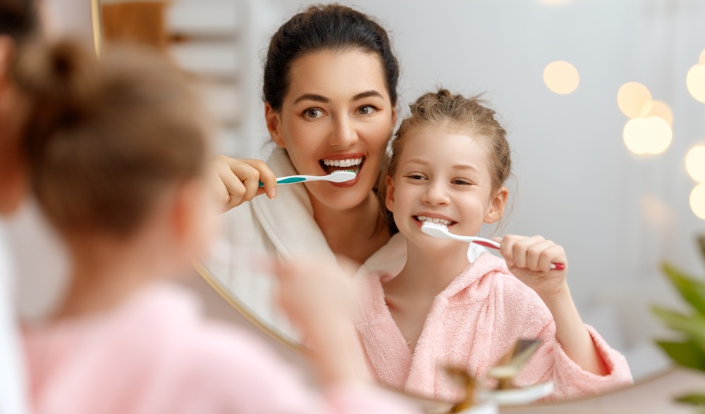 A mother and daughter in pink robes smile as they brush their teeth together in front of a bathroom mirror – Dentist by Union Square