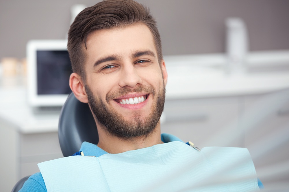 Patient Smiling in Dental Chair – Bone Grafting A young man with a beard smiles while sitting in a dental chair, wearing a blue protective bib, in a clinical setting – Bone Grafting