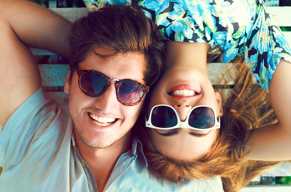 A smiling couple lies on a bench with their heads together, wearing sunglasses and colorful clothes, enjoying a carefree moment – Emergency Dentist East Village