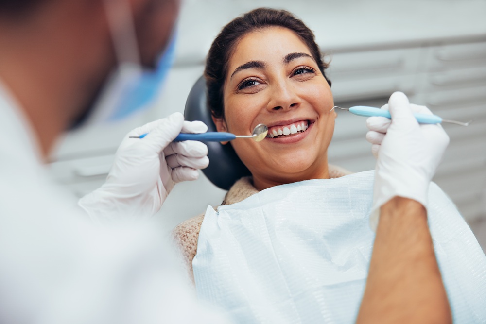 A woman sitting in a dental chair smiling as a dentist performs an oral examination using dental tools – Dentist Gramercy Park NYC