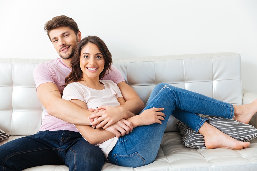 A cheerful couple relaxes on a white leather couch, sitting closely together and smiling at the camera – Emergency Dentist East Village