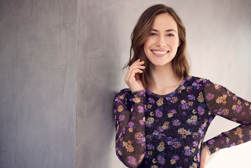 A woman in a sheer floral long-sleeve top stands smiling against a textured gray wall – Chelsea Dentist