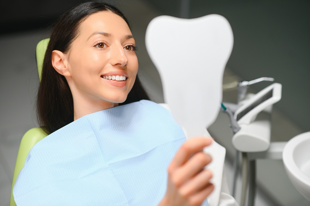 Patient Smiling at Mirror – Dental Implants Gramercy Park A woman with a dental bib smiles while holding a tooth-shaped mirror in a dental chair – Dental Implants Gramercy Park