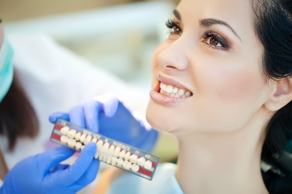 Smile Assessment – Crowns & Bridges A woman smiles during a dental consultation as a gloved hand holds a shade guide to match her teeth color, typically used for crowns and bridges procedures - Crowns & Bridges