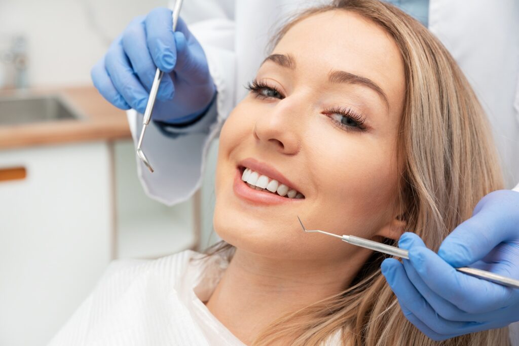 A smiling woman reclines in a dental chair as a dentist wearing gloves and a mask examines her teeth with a mirror and dental probe. - Gramercy Dental Group