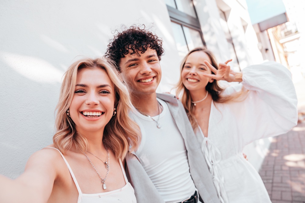 A group of three young friends, dressed in stylish outfits, smile brightly while taking a selfie outdoors on a sunny day. - Gramercy Dental Group 
