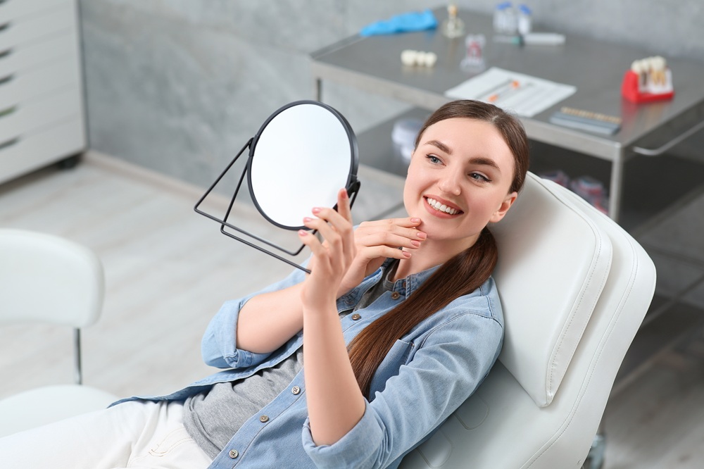 Happy Patient with Mirror – Dental Implants Gramercy Park A smiling young woman reclines in a dental chair, admiring her reflection in a handheld mirror after treatment – Dental Implants Gramercy Park