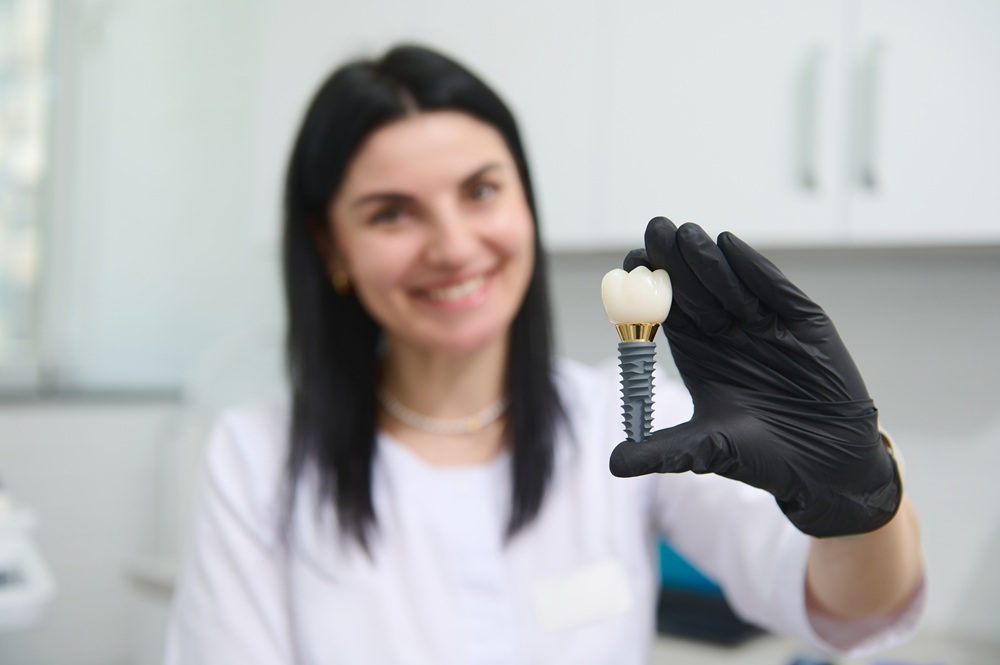 Dentist Holding Dental Implant Model – Chelsea Dental Implants A smiling dentist wearing black gloves holds up a dental implant model in a bright clinical setting – Chelsea Dental Implants