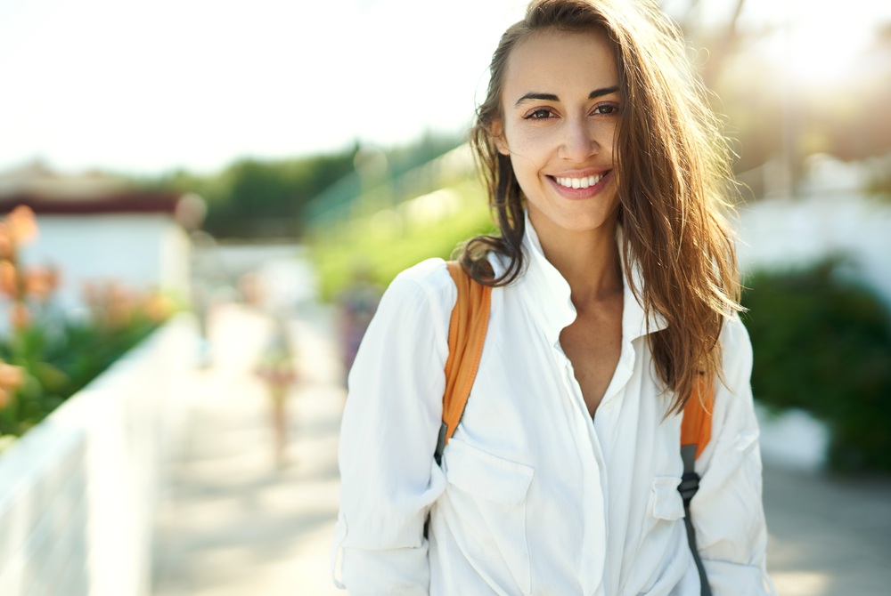 A woman with long brown hair wearing a white shirt and orange backpack straps smiles outdoors in bright sunlight – Chelsea Dentist